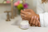 Lavender Shea Butter Body Cream jar placed on a bathroom countertop with hands applying lotion.