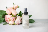 Soul Touch Massaging Set spray bottle next to pink peonies, perfect for skincare and self-care routines.
