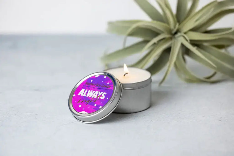 Lavender scented candle in a tin with the label "ALWAYS" next to a decorative plant on a light background.